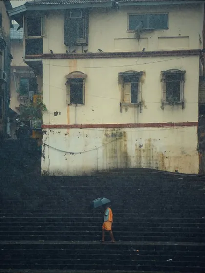 A reprint of the Banganga steps photo, this time as part of the "Stories Everywhere" series. A lone figure in orange walks with an umbrella, creating a solitary mood against the old, weathered building.