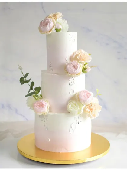 A classic three-tiered wedding cake with a subtle pink ombre effect on the bottom tier. The cake is decorated with silver leaf and clusters of beautiful fresh flowers for a timeless and romantic feel.