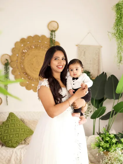 A mother holding her baby boy, both dressed in elegant white outfits. The boho-chic background adds a touch of style to this family portrait.