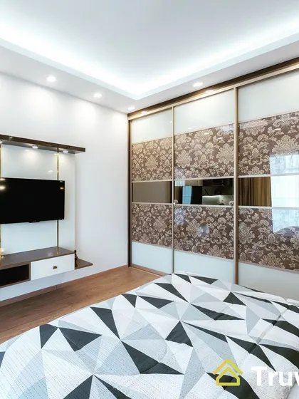 A guest bedroom featuring a sliding wardrobe with decorative fabric panels and mirrors. The design also includes a wall-mounted TV unit for entertainment.