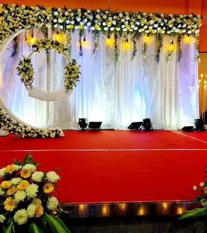 A wide view of a naming ceremony stage inside a hall. The floral moon arch is complemented by elegant white curtains and warm lighting, creating a serene and beautiful ambiance for the event.