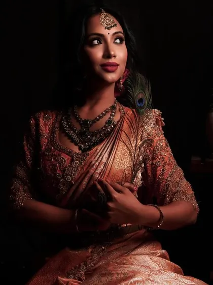 A dramatic and moody shot from a Deepavali-themed shoot. The makeup is rich and warm, with bronze tones and a deep red lip, complementing the ornate saree and antique-finish jewellery.