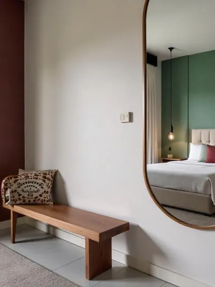 A refined mix of warm and cool tones, this bedroom view shows a large oval mirror reflecting the bed, with a simple wooden bench and a rich burgundy accent wall adding depth.