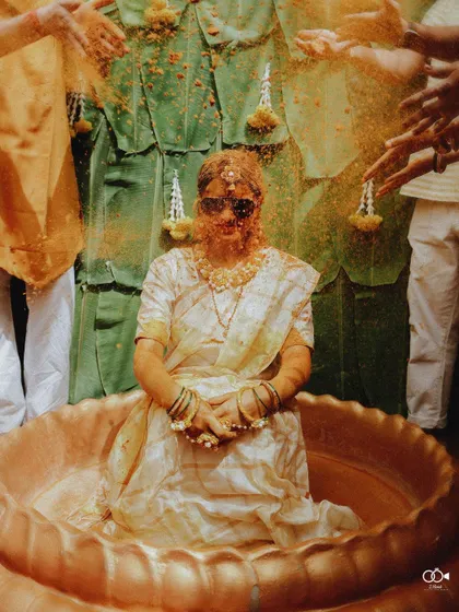 A fun shot of the bride in a brass vessel, covered in haldi and flower petals, wearing sunglasses.