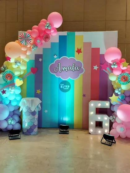 A colorful rainbow-themed backdrop with a lit-up number '6' and whimsical balloon clouds.