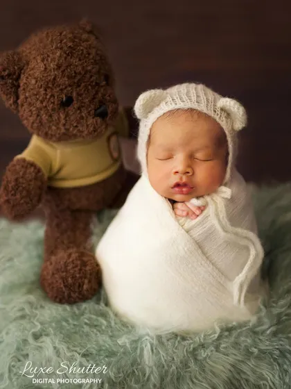 A simple and sweet newborn portrait. The baby, wearing a knitted bear-ear bonnet, is swaddled next to a classic brown teddy bear.