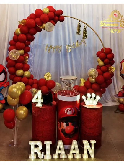 A dynamic superhero-themed birthday decoration featuring characters like Spiderman and Iron Man. The setup uses red and gold balloons on a ring frame and themed plinths.