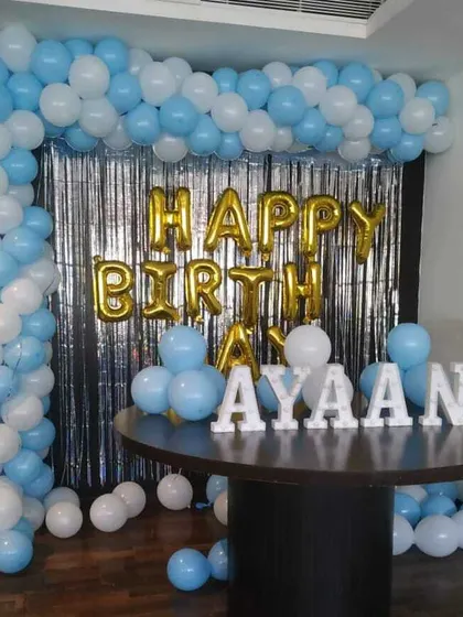 A simple birthday corner setup with a blue and white balloon arch against a silver fringe curtain. Foil 'Happy Birthday' balloons and marquee letters for the name are placed on a table.