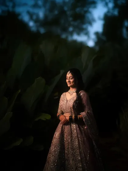 A bride in her reception lehenga, surrounded by the soft darkness of a garden at night. Her gentle smile and the elegant setting create a romantic, fairytale mood.