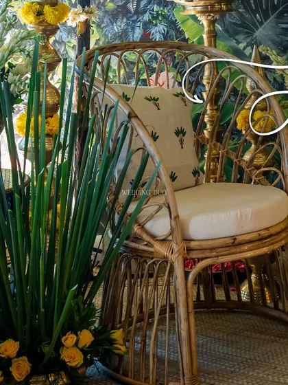 A cozy rattan chair set against a tropical print backdrop. The combination of natural textures, lush greenery, and marigold flowers creates an earthy and lively atmosphere for a Haldi celebration.