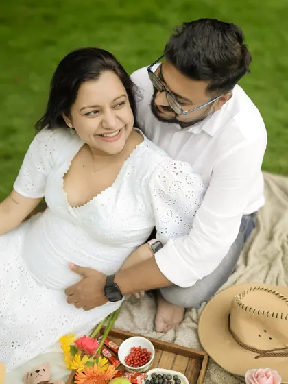 A joyful moment during an outdoor picnic maternity shoot. The couple shares a happy, loving look, surrounded by the fresh greenery of a park.
