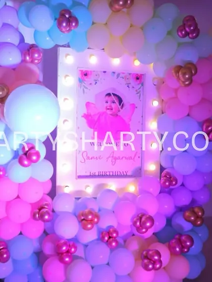 A personalized welcome sign for a first birthday, framed with lights and a dense arrangement of pastel balloons, creating a beautiful photo spot.