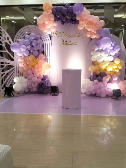 A stunning butterfly-themed first birthday stage in shades of lavender and peach. The decor features large butterfly wing cutouts, a light-up number '1', and beautiful balloon cascades, creating a dreamy and elegant backdrop.