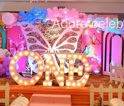 A complete butterfly-themed first birthday stage. The setup includes "ONE" marquee letters, a white picket fence, a photo frame, and butterfly wing props against a sequin wall.