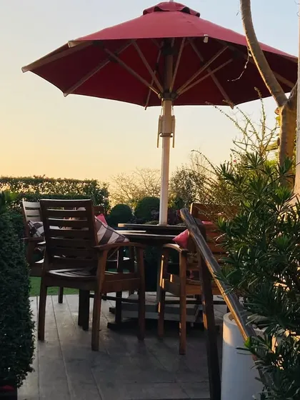 A view of the terrace dining area at sunset, with the red umbrella providing a pop of color. The surrounding greenery and warm light create a perfect setting for an evening outdoors.