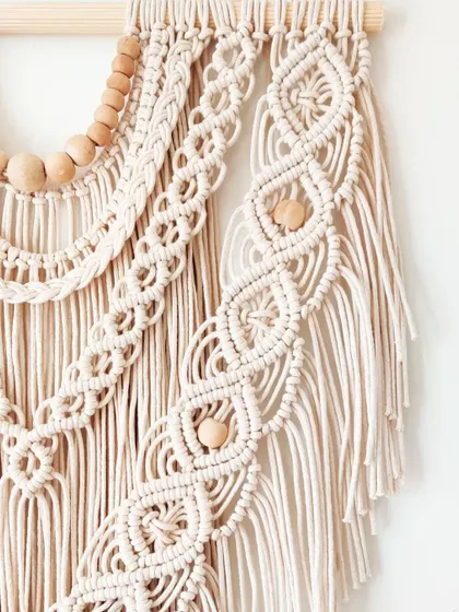 A close-up of a custom wall hanging in a natural off-white color, showing the detailed knot work and wooden bead accents.