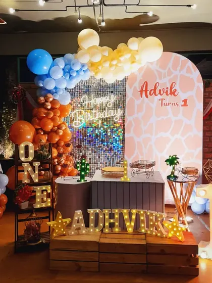 A vibrant first birthday setup for Advik with a giraffe-print arch and a shimmering backdrop. The combination of orange, blue, and yellow balloons adds a playful pop of color.