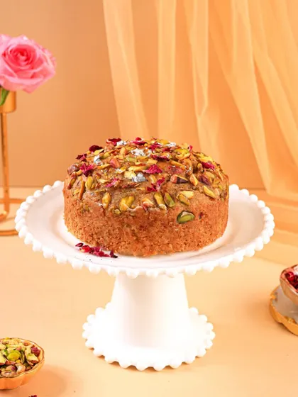 A beautiful Mawa cake topped with pistachios and rose petals, presented on a classic white cake stand for an elegant look.