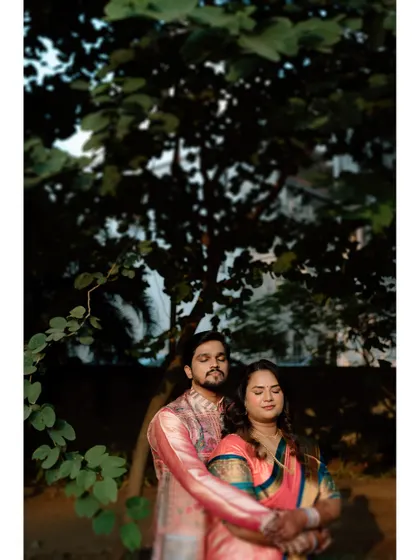 A moody and artistic portrait of a couple embracing under a large tree at dusk. The low light creates a romantic and intimate atmosphere.
