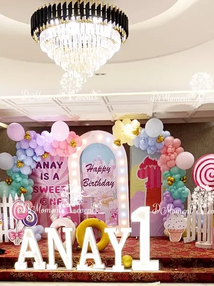 A sweet and imaginative Candyland stage for a first birthday, featuring giant ice cream and lollipop props, a white picket fence, and pastel balloon arrangements.
