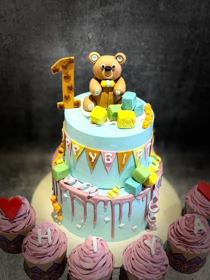 A sweet two-tier teddy bear cake for a first birthday, complete with matching cupcakes. The design is simple, cute, and perfect for celebrating a little one's milestone.