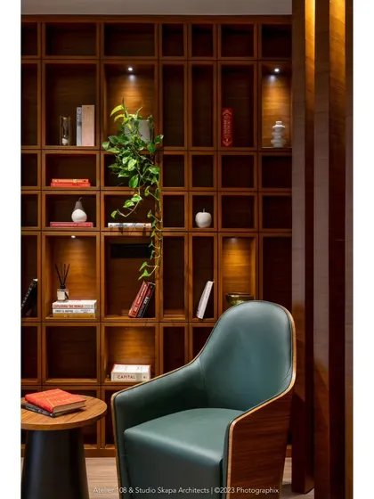 A cozy reading nook in the Meghana Foods office, featuring a plush green chair set against a wall of rich wooden shelves. The shelves are designed with assorted sizes and are individually lit to highlight decorative objects and books.