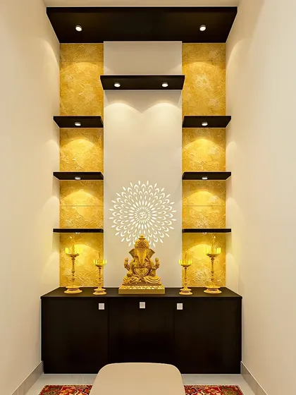 A tall pooja unit with dramatic backlighting on a textured gold wall. The multiple black shelves provide a striking contrast and plenty of space for idols.