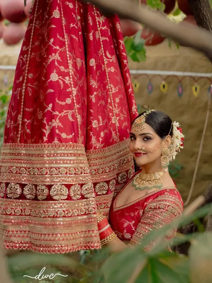 A beautiful bridal portrait, with the bride peeking out from behind her stunning red wedding lehenga, her smile full of anticipation.