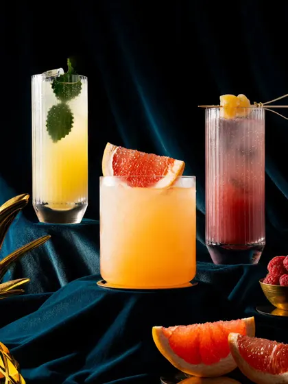 A trio of colorful cocktails for Nara Thai, shot against a deep blue velvet curtain. The jewel tones of the drinks and the rich fabric create a luxurious and elegant mood.