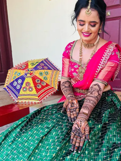 A vibrant bride in a pink and green lehenga, her hands covered in beautiful, dark-stained mehendi featuring traditional bride and groom figures.