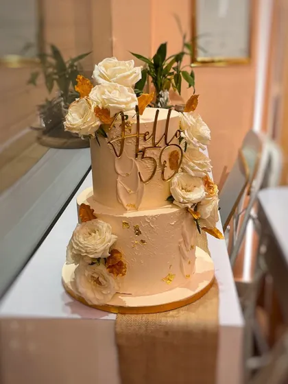 "Hello 50" has never looked so good. This two-tier cake features a textured buttercream finish, adorned with an abundance of fresh white roses and gold leaf details, making it perfect for a milestone celebration or an elegant wedding.