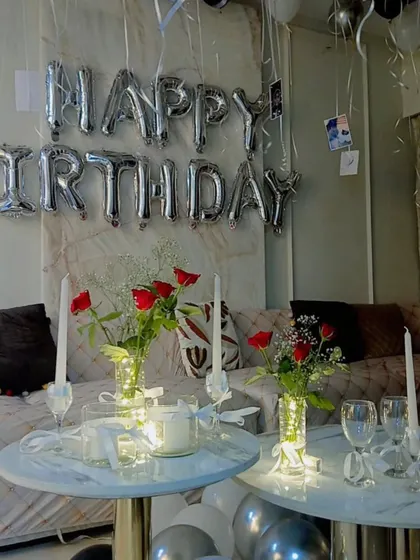 An elegant and simple birthday surprise setup at home. We decorated the living room with silver 'Happy Birthday' balloons and set up tables with candles, red roses, and fairy lights.
