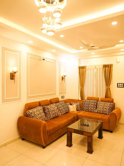 A warm and inviting living room designed with a comfortable tan sectional sofa and classic wall molding. The elegant chandelier and recessed ceiling lights provide a soft, ambient glow throughout the space.