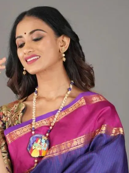 A model showcases a hand-painted pichwai-style pendant necklace depicting Radha and Krishna. This artistic piece combines traditional painting with jewellery making for a unique accessory.