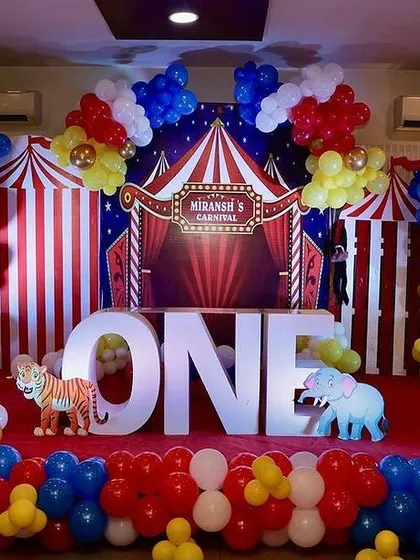 A carnival or circus themed first birthday stage. The decoration features a red and white striped backdrop, colorful balloon arrangements, and large marquee letters spelling "ONE".