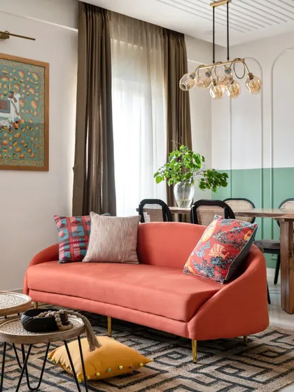 The vibrant orange sofa is the captivating centerpiece of this living room. Set against a backdrop of dual-toned walls and nested cane tables, it infuses the space with energy and personality.