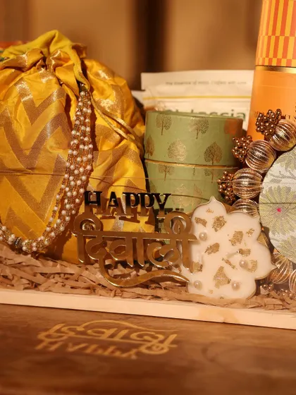 A 'Happy Diwali' acrylic cutout placed inside a hamper, alongside a pearl-string adorned potli and a floral candle.