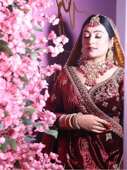 A serene bridal portrait with a backdrop of pink blossoms. The bride wears a classic maroon velvet lehenga, her look softened by the floral setting.