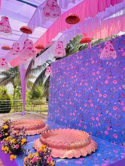 The seating for the couple at the purple-themed Haldi, featuring a custom floral printed backdrop and lotus-shaped brass urlis.