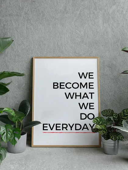 "We Become What We Do Everyday." This powerful motivational quote, shown here in a natural wood frame amongst house plants, serves as a simple yet profound daily reminder of the power of habits.