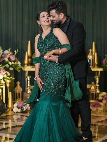 A full-length portrait showcasing the couple's stunning cocktail night attire. The bride's mermaid-style green gown and the groom's all-black suit create a look of modern sophistication.