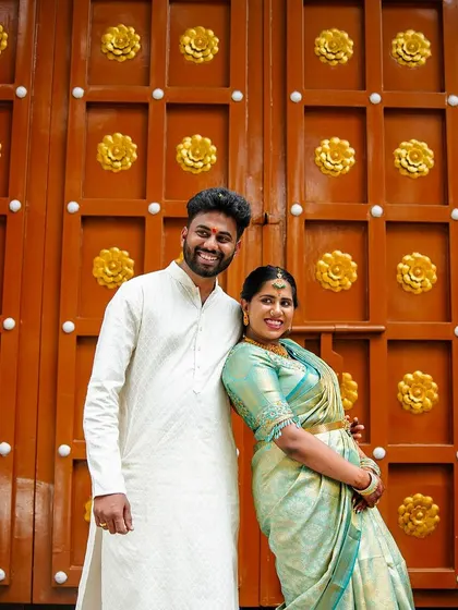 A happy, relaxed portrait of an engaged couple in front of a grand, golden temple door. Their coordinated outfits and easy smiles show their excitement.