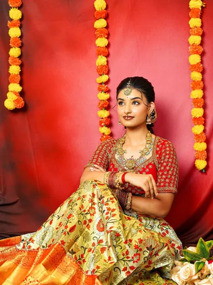 A full view of a bride-to-be in a traditional half-saree, adorned with an antique jewellery set. The red background makes the gold and green of the jewellery pop.