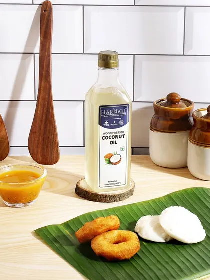 This shot places the coconut oil in a South Indian kitchen context, styled with idli, vada, and traditional wooden spoons. It effectively communicates a key use case for the product in regional cooking.