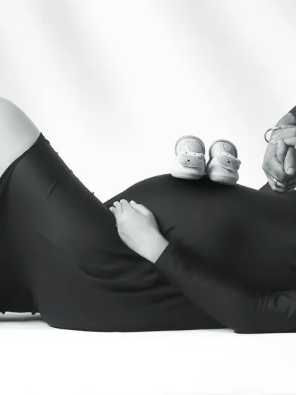 A creative black and white shot focusing on the details. A pair of baby shoes rests on the mother's belly as she lies down, her partner's hand gently holding hers.
