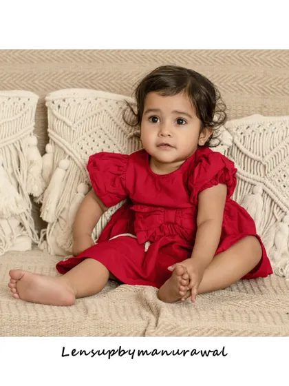 A charming portrait of a toddler in a red dress, looking inquisitively to the side while sitting on a comfortable, modern sofa in the studio.
