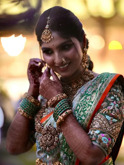 A bride getting ready for her big day. Her green and orange saree is paired with a heavily embroidered blouse featuring intricate bird and floral motifs.