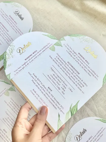 A closer look at a heart-shaped wedding details card. This custom die-cut shape, combined with delicate greenery illustrations and elegant gold foil lettering, adds a touch of romance and whimsy to the invitation suite.