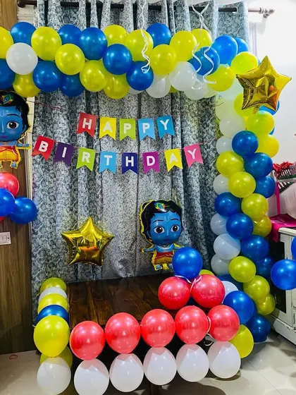 A colorful Krishna-themed birthday party at home. The decoration includes a balloon arch in front of a curtain, with Krishna character foil balloons.