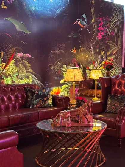 A luxurious lounge corner from the "Jungle" themed party. A classic leather chesterfield sofa is set against a vibrant, tropical-print wallpaper, creating a moody and sophisticated vibe.